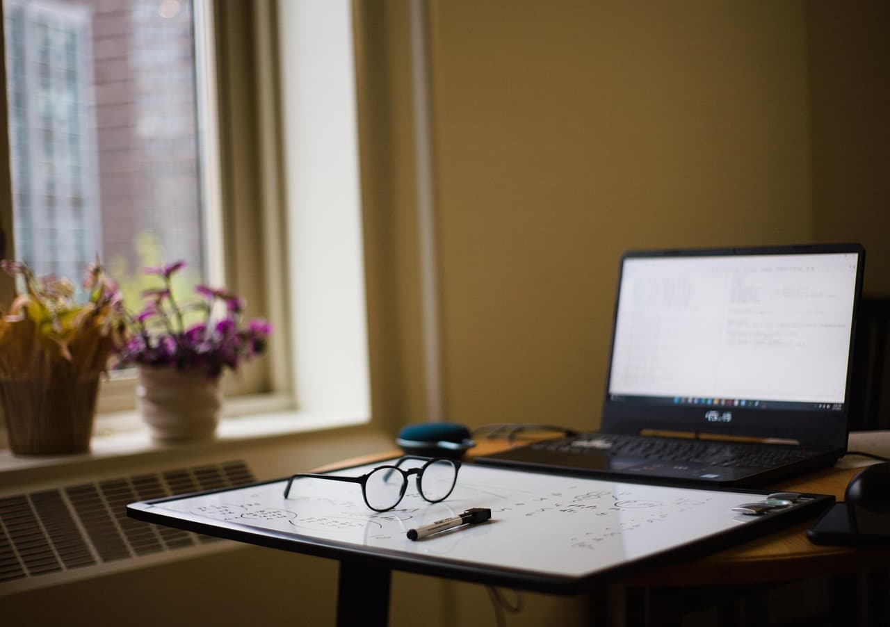 Ein Laptop offen auf einem Schreibtisch am Fenster, daneben ein Notizblock, ein Stift und eine Brille