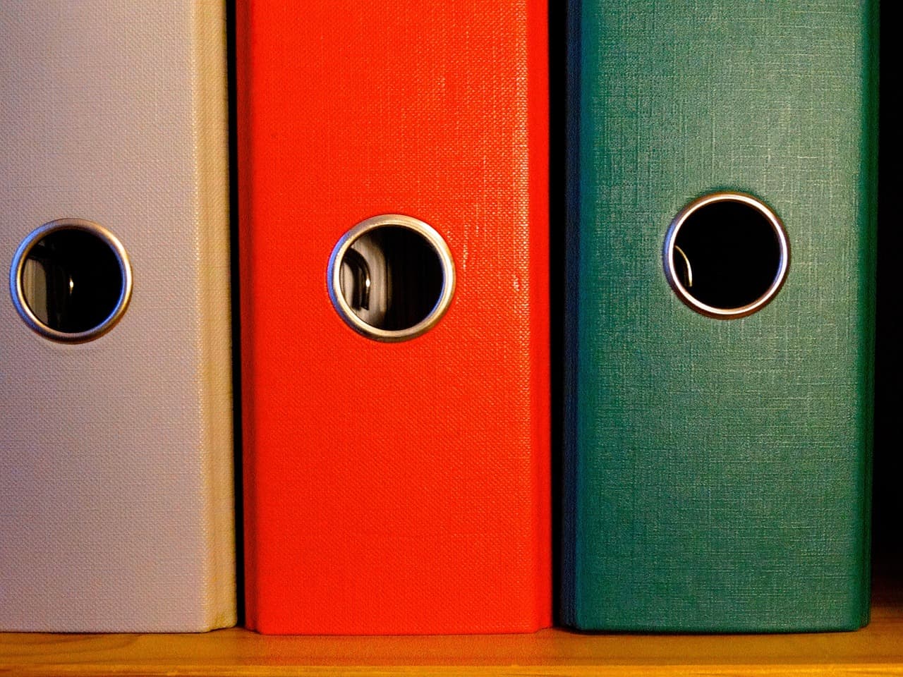 Three lever-arch binders side by side on a shelf, in grey, red and green, each with a metal finger hole
