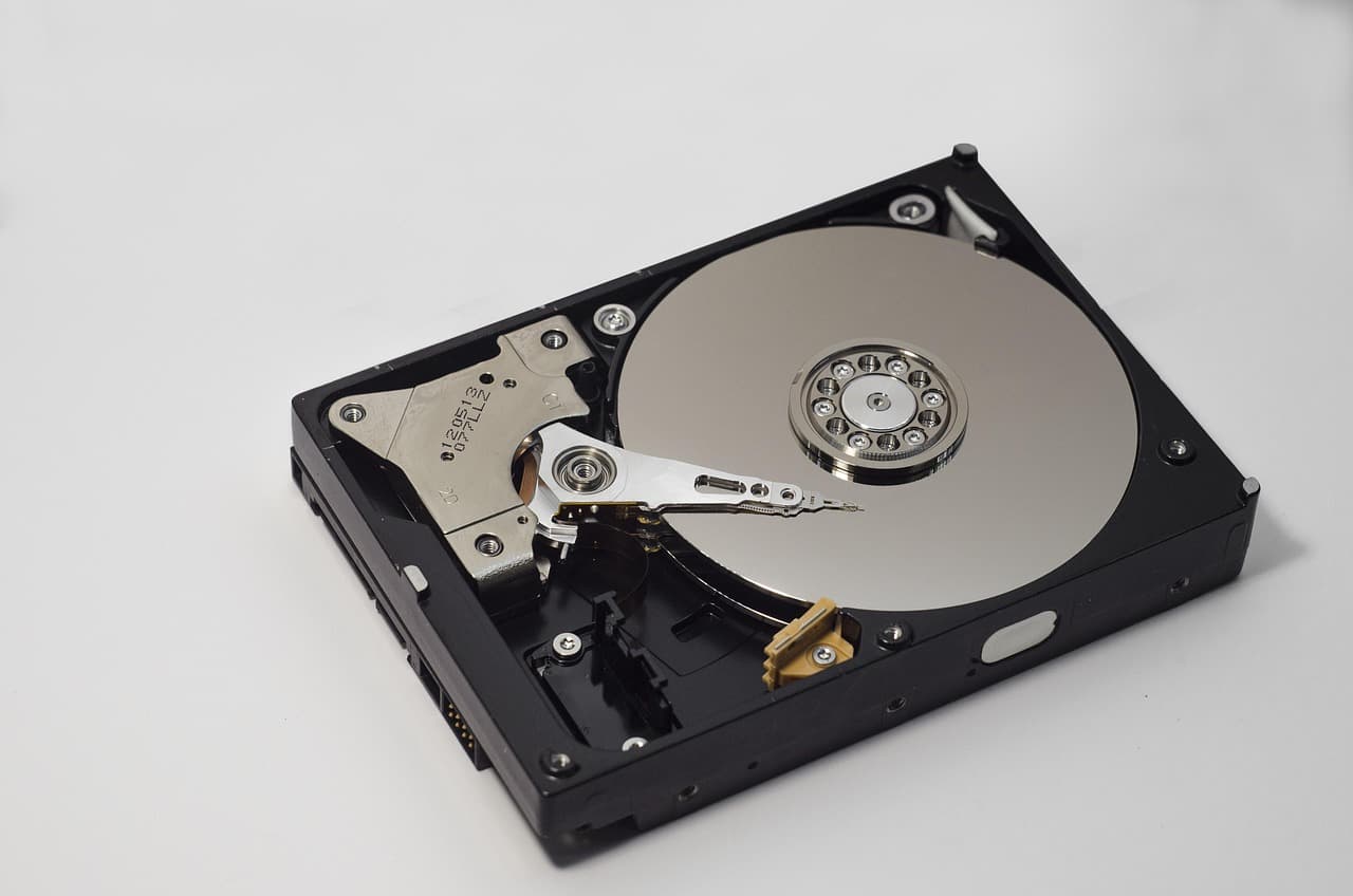 Um disco rígido de 3,5 polegadas com a tampa retirada sobre fundo branco, deixando ver o prato com acabamento de espelho, a cabeça de leitura pousada no seu braço por cima do disco e o cubo aparafusado